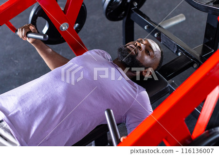 An African American young male athlete, a fitness model lying on a bench in a gym, lifting weights An African American young male athlete, a fitness model lying on a bench in a gym, lifting weights 116136007