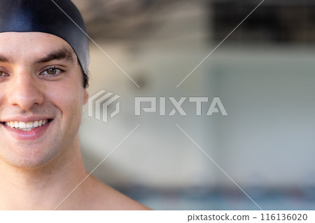 Caucasian young male swimmer standing indoors, wearing black swim cap, smiling, copy space 116136020
