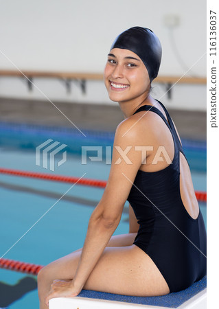 A biracial young female swimmer sitting poolside indoors, smiling at the camera, copy space A biracial young female swimmer sitting poolside indoors, smiling at the camera, copy space 116136037