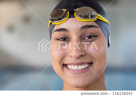 Young biracial female swimmer wearing goggles and cap, smiling at the camera indoors 116136041