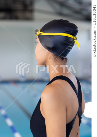 Young biracial female swimmer wearing a black swimsuit and cap, standing poolside indoors Young biracial female swimmer wearing a black swimsuit and cap, standing poolside indoors 116136043