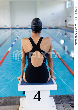 Young biracial female swimmer sitting on starting block at an indoor pool Young biracial female swimmer sitting on starting block at an indoor pool 116136056