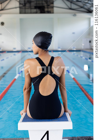Biracial young female swimmer sitting on starting block indoors, looking at pool Biracial young female swimmer sitting on starting block indoors, looking at pool 116136061