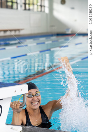 Biracial young female swimmer wearing goggles celebrating indoors in pool, copy space 116136089