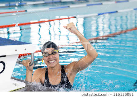 Biracial young female swimmer celebrating indoors in pool, wearing goggles 116136091