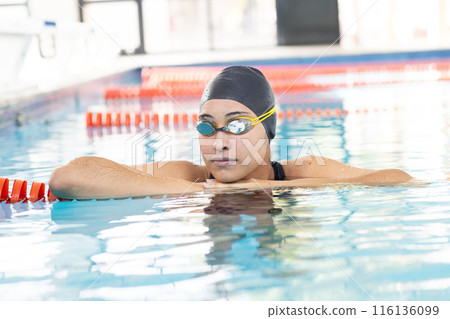 Young biracial female swimmer resting at pool edge indoors, wearing goggles and swim cap 116136099