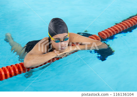 Biracial young female swimmer training indoors in pool, wearing black cap and goggles Biracial young female swimmer training indoors in pool, wearing black cap and goggles 116136104