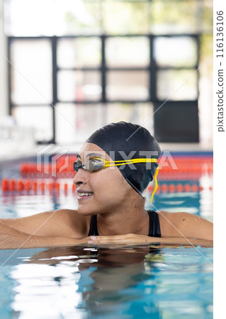 Young biracial female swimmer resting at pool edge indoors, wearing goggles Young biracial female swimmer resting at pool edge indoors, wearing goggles 116136106