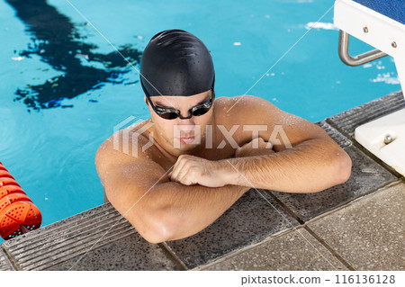 Caucasian young male swimmer resting at pool edge indoors, wearing goggles Caucasian young male swimmer resting at pool edge indoors, wearing goggles 116136128