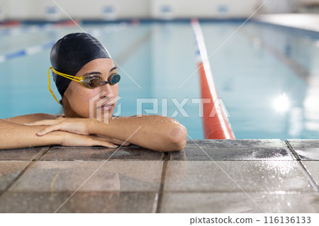 A biracial young female swimmer resting at poolside indoors, wearing goggles, copy space 116136133