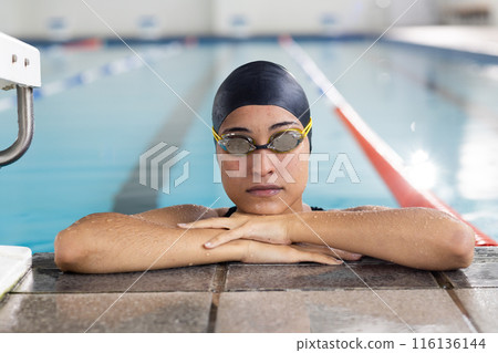 Young biracial female swimmer resting at pool edge indoors, wearing goggles 116136144