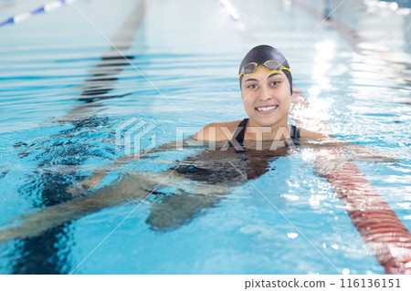 Biracial young female swimmer resting at pool edge indoors, wearing goggles Biracial young female swimmer resting at pool edge indoors, wearing goggles 116136151