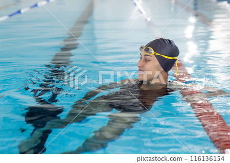 Young biracial female swimmer resting at pool edge indoors, wearing goggles 116136154