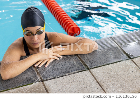 A biracial young female swimmer resting at poolside indoors, wearing goggles A biracial young female swimmer resting at poolside indoors, wearing goggles 116136156