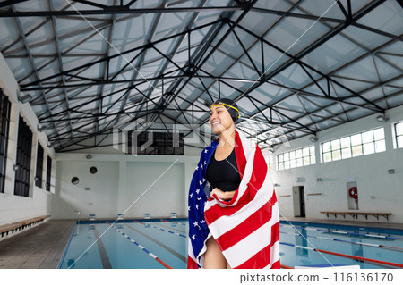 Young biracial female swimmer standing by pool indoors, wrapped in American flag, copy space 116136170