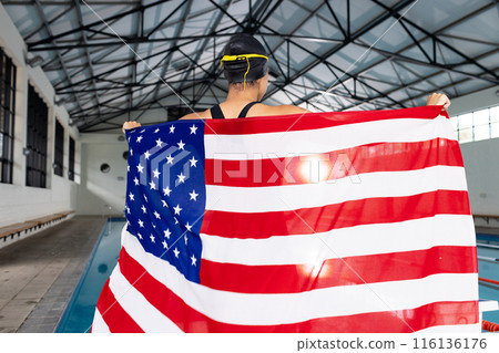 Young biracial female swimmer holding American flag by poolside indoors Young biracial female swimmer holding American flag by poolside indoors 116136176