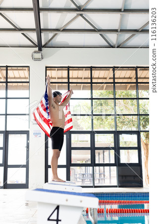 Caucasian young male swimmer holding American flag, standing on poolside indoors Caucasian young male swimmer holding American flag, standing on poolside indoors 116136203