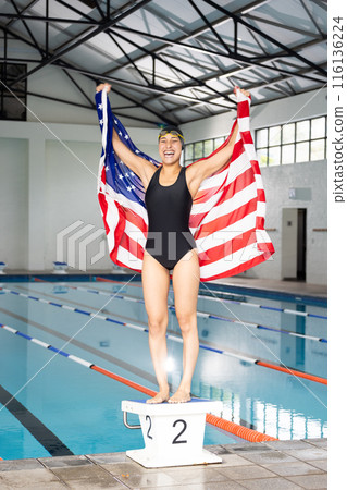 Young biracial female swimmer holding American flag, standing by pool indoors 116136224