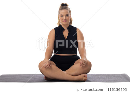 A young Caucasian plus-size model sits cross-legged on yoga mat, white background, copy space A young Caucasian plus-size model sits cross-legged on yoga mat, white background, copy space 116136593
