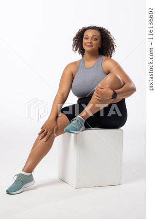 A biracial young female plus size model sits on white block, white background, copy space 116136602