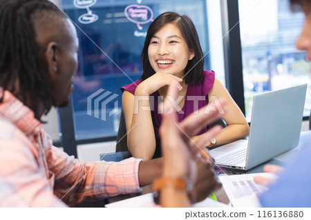 Diverse team discussing around laptop in a modern business office, Asian woman smiling 116136880