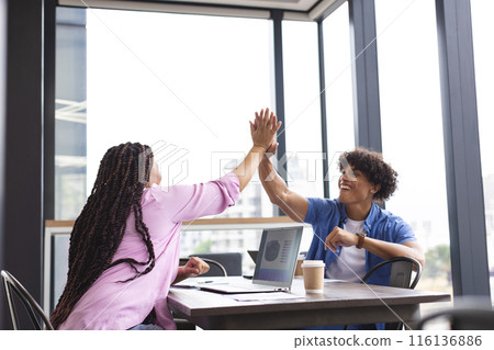 Diverse colleagues high-fiving at table with a laptop in a modern business office Diverse colleagues high-fiving at table with a laptop in a modern business office 116136886