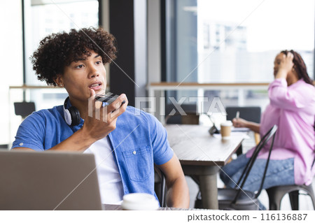 Biracial man with headphones talks on a smartphone, woman nearby in modern office 116136887