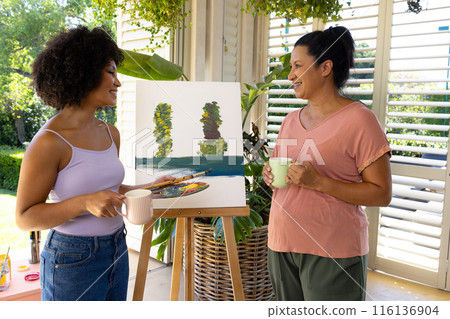 Mature biracial mom with coffee admires painting by young biracial woman Mature biracial mom with coffee admires painting by young biracial woman 116136904