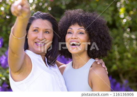 Mature biracial mother points, young daughter smiles beside her in garden 116136906