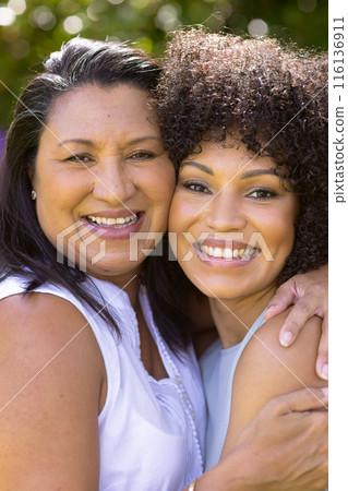 Mature biracial mother embraces her young daughter, both smiling in the garden 116136911