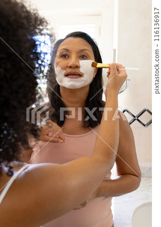 Mature biracial woman and daughter use facial masks, looking in mirror 116136917