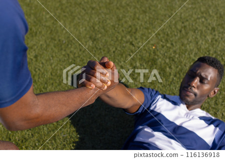 Two African American male athletes train on a field, one assists the other to stand 116136918