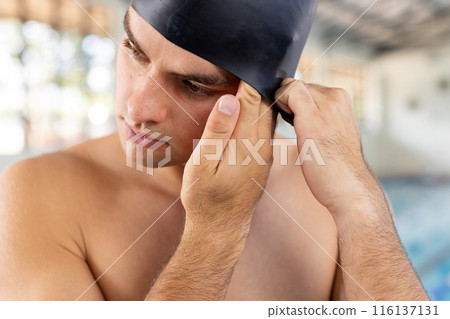 Caucasian young male swimmer adjusting black swim cap indoors, standing by pool Caucasian young male swimmer adjusting black swim cap indoors, standing by pool 116137131