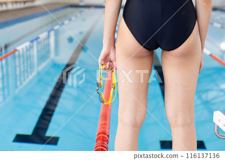 Biracial young female swimmer holding goggles stands by pool lane indoors, copy space 116137136