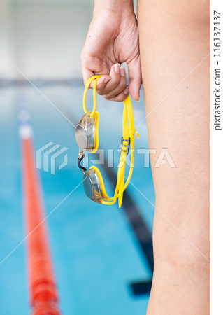 A biracial young female swimmer holding yellow goggles stands poolside indoors 116137137