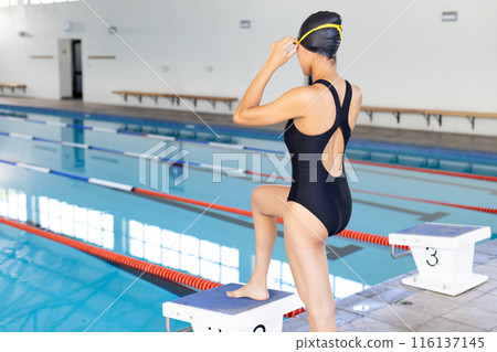 Biracial young female swimmer adjusting goggles at poolside indoors, copy space Biracial young female swimmer adjusting goggles at poolside indoors, copy space 116137145