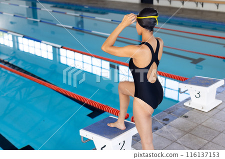 Biracial young female swimmer standing on starting block indoors, looking forward, copy space 116137153