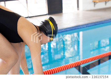 Biracial young female swimmer preparing to dive into an indoor pool, copy space 116137154