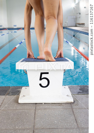 Biracial young female swimmer standing on starting block indoors, ready to dive 116137167