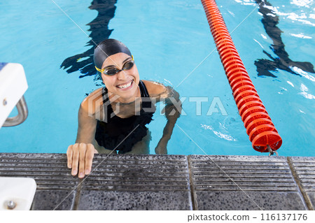 Biracial young female swimmer is hanging onto the pool edge indoors, smiling 116137176
