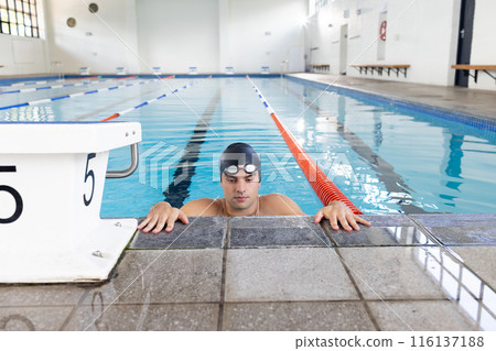 Caucasian young male swimmer wearing cap and goggles, resting at swimming pool edge 116137188