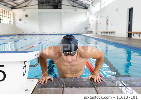 Caucasian young male swimmer resting at swimming pool edge indoors, wearing goggles 116137191