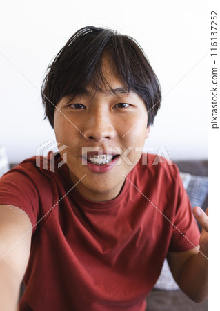 Asian teen boy with braces takes a selfie and video calls at home in a red shirt Asian teen boy with braces takes a selfie and video calls at home in a red shirt 116137252