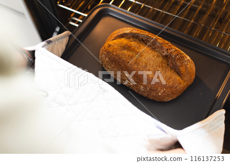 Hands wearing oven mitts taking out freshly baked bread from oven at home with copy space 116137253