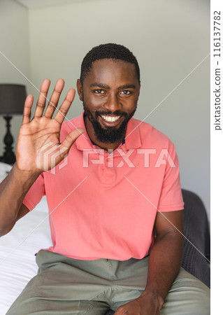 African American man waving during a video call, sitting on bed at home 116137782