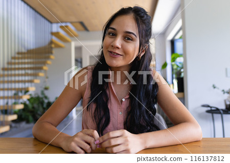 Young biracial woman sitting at table, smiling at camera on a video call at home 116137812