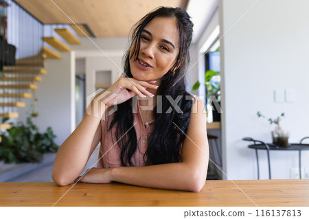 Biracial young woman sitting at table, resting chin on hand, smiling at home on a video call 116137813