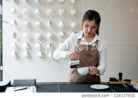 An experienced Asian female barista pouring milk into a coffee cup, working in a coffee shop. 116137910