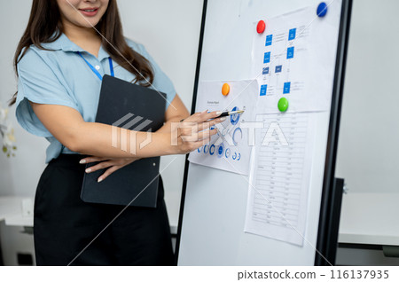 An Asian businesswoman is standing in the meeting, pointing her pen at a paper on the whiteboard. 116137935