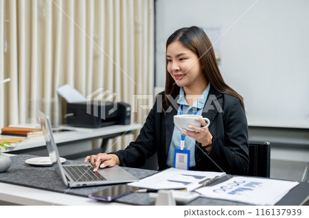 A happy Asian businesswoman is having coffee while working on her laptop computer at her desk. A happy Asian businesswoman is having coffee while working on her laptop computer at her desk. 116137939